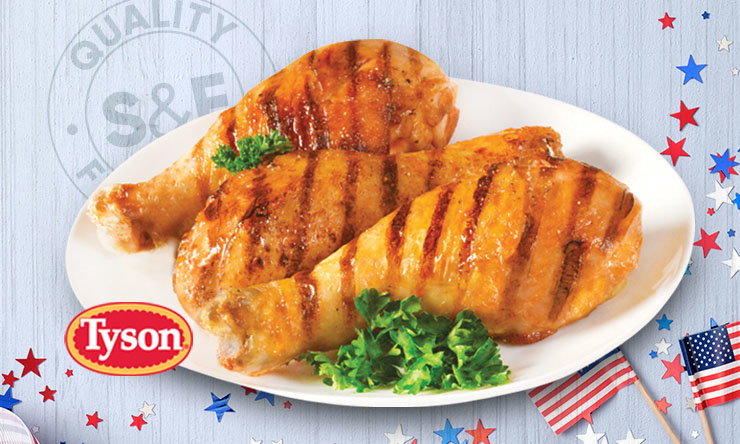 A white plate holding three grilled Tyson chicken drumsticks with visible grill marks, garnished with parsley, placed on a light wood background decorated with red, white, and blue stars and small American flags